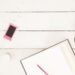Flat lay of an open notebook with a pink pencil, coffee cup lid, and pink flower petals on a white wooden table — feminine workspace concept.