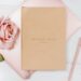 Flat lay of a beige “Daily Goal Setter” notebook on top of a pink laptop with a pink rose, pastel pen, paper clips, and a folded fan on a white wooden surface — elegant and feminine workspace setup.