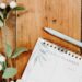A flat lay of a weekly planner and light blue pen on a wooden surface, accented with green leaves and white berries for a natural, cozy look.