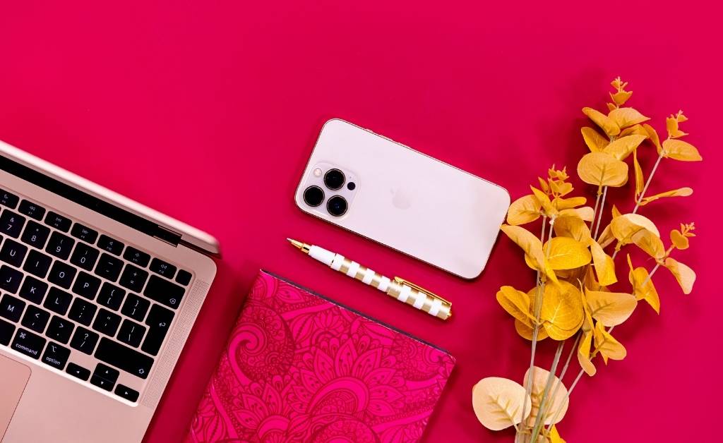 A flat lay of a workspace on a bright pink background featuring a laptop, smartphone, patterned notebook, elegant pen, and a small branch with yellow leaves.