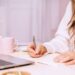 Person writing a short plan in a notebook beside a laptop and coffee, representing small, focused steps to move forward in business.