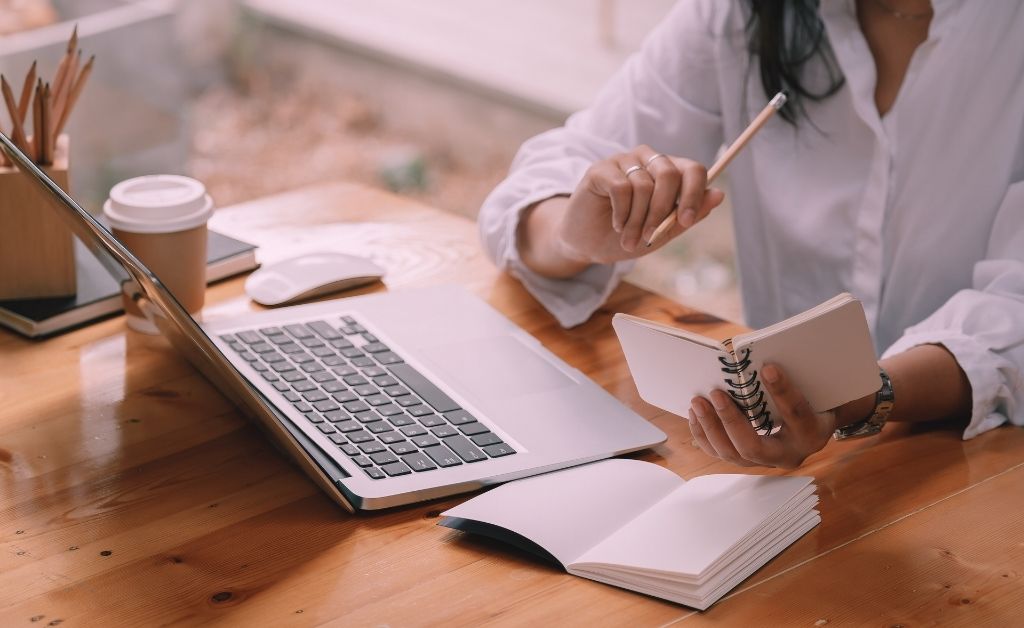 Person sitting at a wooden desk with an open laptop, writing in a small notebook beside a larger open journal and a cup of coffee.