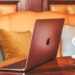 Rose-gold laptop on a wooden café table with two cups of coffee in front of a cozy couch with colorful pillows.