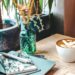 Latte with swan latte art on a wooden café table beside a notebook, pen, laptop, and a small green glass vase with dried flowers.