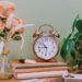 Soft, minimalist workspace with a vintage alarm clock resting on stacked books, blush roses in a glass vase, and a leafy green plant against a calm green wall, reflecting quiet presence and intentional rhythm.