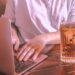 Person typing on a laptop at a wooden café table beside a glass of iced coffee, representing focused, thoughtful work on building a meaningful business offer.