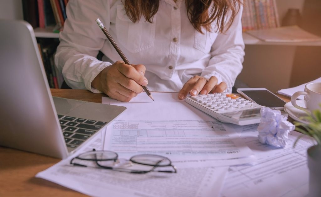 A person in a white shirt sitting at a wooden desk, focused on writing notes amidst a clutter of business tools including a laptop, calculator, tax forms, and a crumpled piece of paper. The warm lighting captures the "smart and soulful" tension of working through business roadblocks.