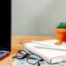 A cozy wooden desk setup featuring a laptop, a pair of black-rimmed glasses, and two pens resting on a stack of cream and mint green planners, with a small succulent and a monstera leaf in the background.