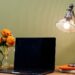 A warm workspace with a laptop centered between a vase of orange roses and a modern mesh desk lamp, set against a sage green wall with a stack of books and a monstera plant.