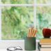 A bright window sill with a stack of books, a shiny red apple, a pair of glasses, and a green fluted cup filled with colored pencils, with a blurred view of green trees outside.