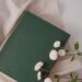 Green hardcover book resting on soft white fabric with small white flowers, symbolizing peace, faith, and quiet reflection