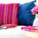 A bright living area featuring a white tufted sofa with two deep blue cushions and one striped pink and orange cushion. An open laptop and pink journals sit on the seat, while a wooden side table to the right holds a stack of books and a vase of vivid pink roses.