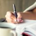 Close-up of a hand writing on lined paper in a bright, soft-lit setting. A white ceramic mug of coffee sits out of focus in the bottom left, creating a peaceful and productive atmosphere.