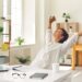 Woman stretching at her desk beside a laptop in a bright home office, representing healthy work habits and building consistent business rhythms without rigid schedules.