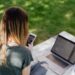 A woman with long blonde hair sitting on a white blanket in a grassy park, working remotely on a laptop and checking her smartphone. High-angle view of a freelancer or student using a mobile phone and computer for digital work in an outdoor setting.