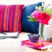 A cozy home office scene on a cream sofa with navy and pink striped pillows, showing a laptop, notepad, and a side table with pink roses, books, and glasses.