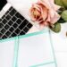 Flat lay of a teal weekly planner and pen resting on an open rose gold laptop keyboard, next to a single pink rose on a white distressed wood background.