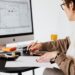 An entrepreneur or architect working at a desk, reviewing paper sketches and an architectural blueprint on a desktop computer monitor, illustrating the concept of designing a business around your life.