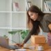 An e-commerce entrepreneur managing an online business, holding shipping boxes while using a laptop at a desk, illustrating the concept of building your own business strategy.