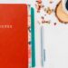 Overhead view of a red notebook with the word NOTES on the cover, a teal journal, a gray pen, and a cup of black coffee on a white desk with dried flower petals.