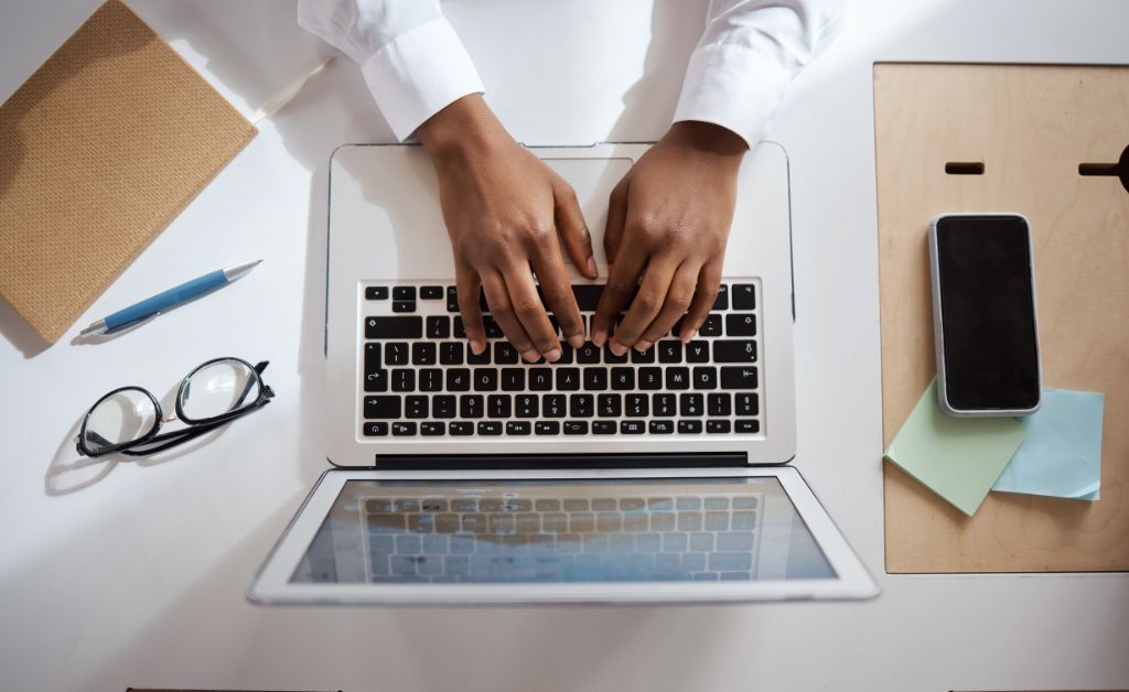 Overhead view of a person typing on a laptop computer keyboard at a clean white desk with a notebook, pen, glasses, smartphone, and sticky notes.