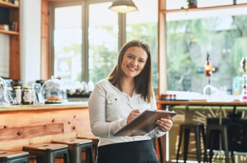 A smiling female server or business owner holding a pen and clipboard inside a brightly lit cafe or restaurant with a wooden counter and bar stools.