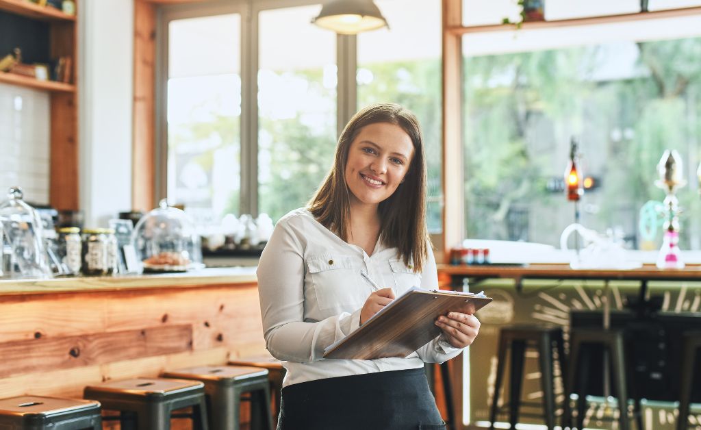 A smiling female server or business owner holding a pen and clipboard inside a brightly lit cafe or restaurant with a wooden counter and bar stools.