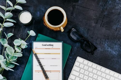 An overhead view of a dark textured workspace featuring a "Goal setting" worksheet, a pen, a cup of black coffee, eyeglasses, a computer keyboard, a small candle, and eucalyptus leaves.