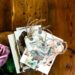 A top-down view of a rustic wooden surface with a pink rose and a bundle of vintage-style postcards and gift tags tied with twine.