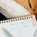 A close-up view of an open spiral-bound weekly planner with a light blue pen resting on it, placed next to a laptop on a wooden surface with a soft, fuzzy white textile.