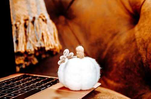 A close-up shot of a small, white decorative fabric pumpkin resting on the corner of a laptop keyboard against a cozy, warm brown background with a fringed blanket.