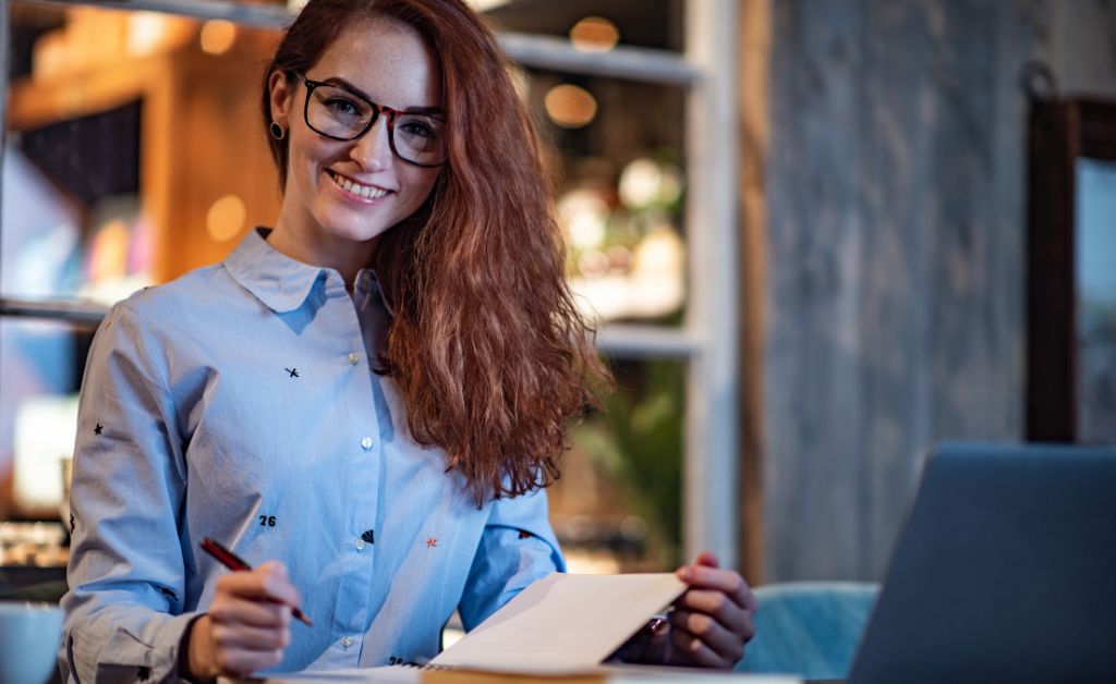 A smiling woman with long wavy reddish hair and glasses sits at a table writing on a notepad with a laptop open nearby in a warmly lit setting.