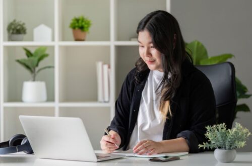 A woman sits at a bright desk writing in a notebook while looking at an open laptop, with potted plants and shelves in the background.