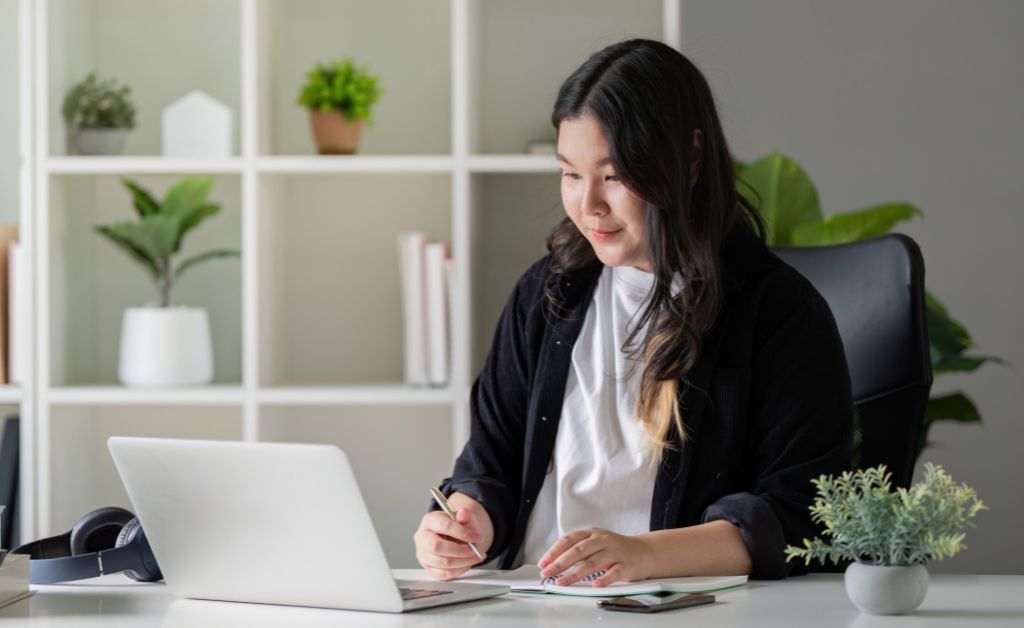 A woman sits at a bright desk writing in a notebook while looking at an open laptop, with potted plants and shelves in the background.