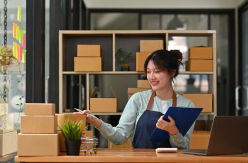 A smiling woman wearing an apron holds a clipboard and points a pen toward stacked cardboard boxes at her desk, with a laptop, calculator, and shelves full of packages in the background.