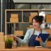 A smiling woman wearing an apron holds a clipboard and points a pen toward stacked cardboard boxes at her desk, with a laptop, calculator, and shelves full of packages in the background.