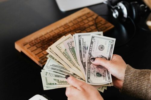 A person counts a fan of United States dollar bills at a desk, with a wooden computer keyboard and headphones in the background.