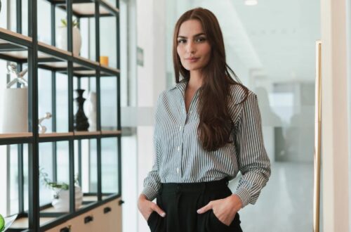 A confident professional woman with long brown hair stands with her hands in her pockets, wearing a striped button-down shirt and black trousers next to a modern office display shelf.