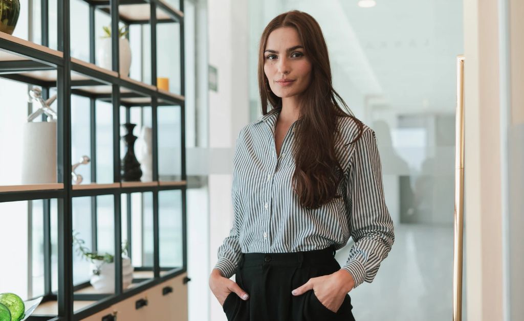 A confident professional woman with long brown hair stands with her hands in her pockets, wearing a striped button-down shirt and black trousers next to a modern office display shelf.