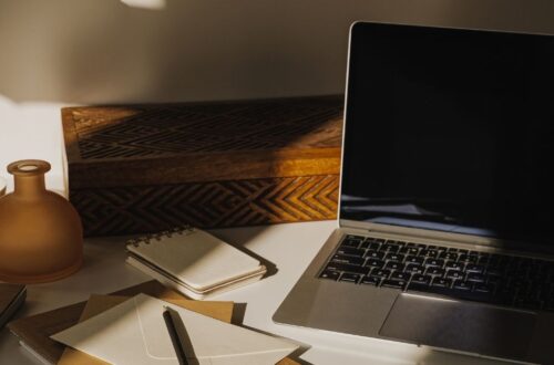 An open laptop sits on a white desk next to stationery envelopes, a small spiral notepad, an amber glass bottle, and a carved wooden box under warm sunlight.