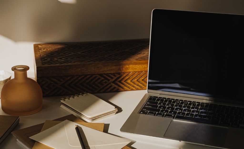 An open laptop sits on a white desk next to stationery envelopes, a small spiral notepad, an amber glass bottle, and a carved wooden box under warm sunlight.
