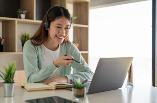 A smiling woman wearing a headset sits at a white table, holding a pen and looking at her open laptop during a video call or online class.