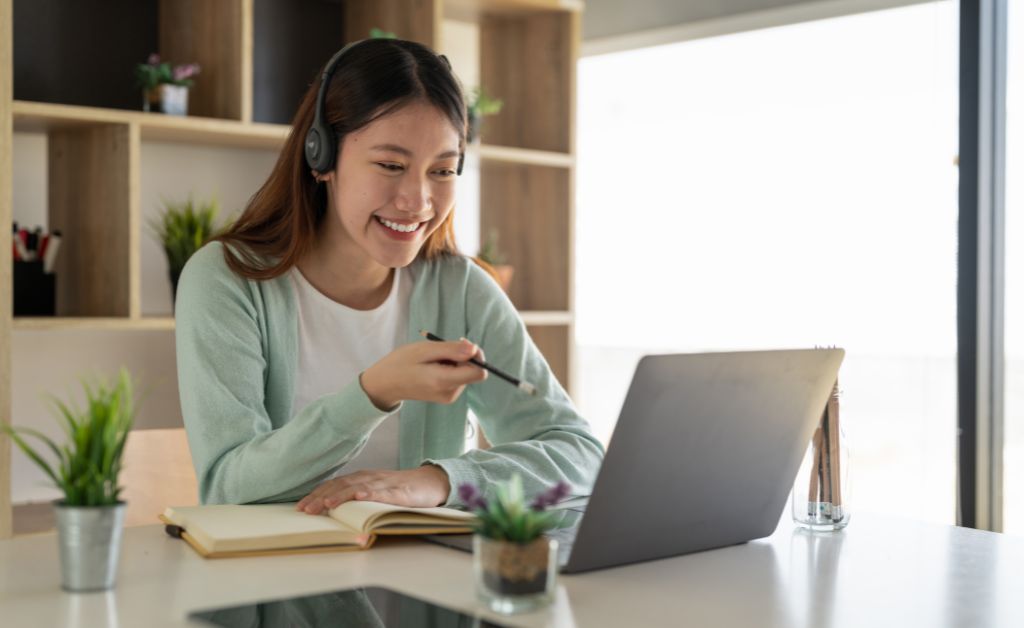 A smiling woman wearing a headset sits at a white table, holding a pen and looking at her open laptop during a video call or online class.