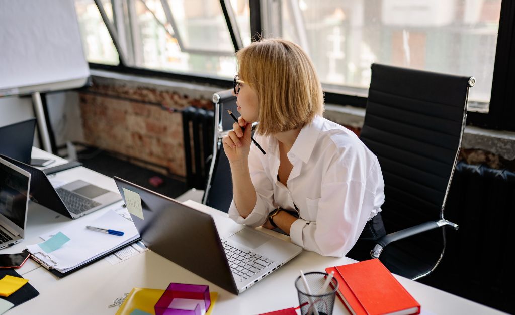 A professional woman with blonde hair wearing a white button-down shirt sits thoughtfully at her office desk, looking away from her open laptop in a bright co-working space.