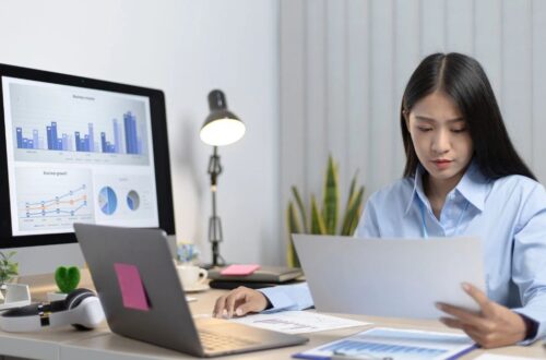 A focused female business analyst reviews printed financial charts at her desk, with business data graphs displayed on a large computer monitor and an open laptop nearby.