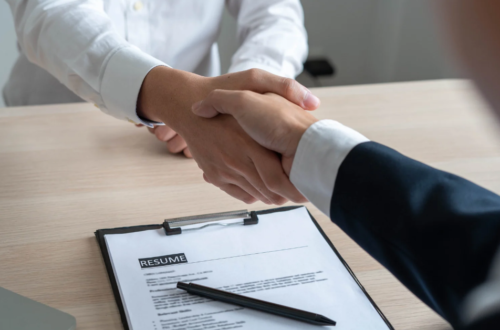 Two professionals shake hands across a desk during a job interview, with a printed resume on a clipboard and a pen visible in the foreground.